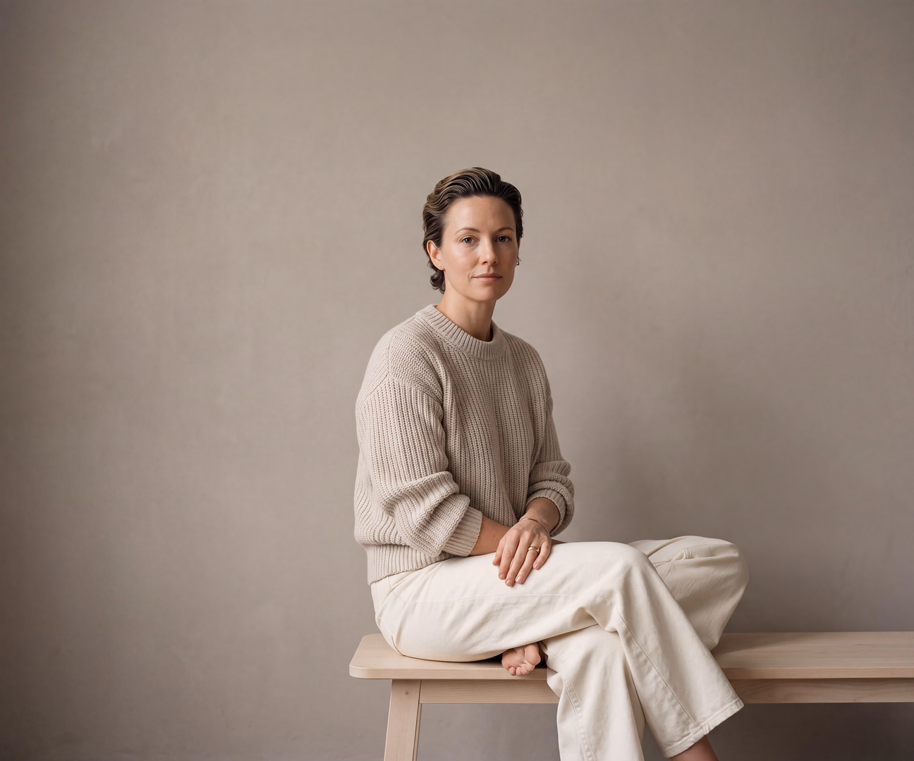 Model in Strickpullover und heller Hose, sitzend vor einer hellen Wand
