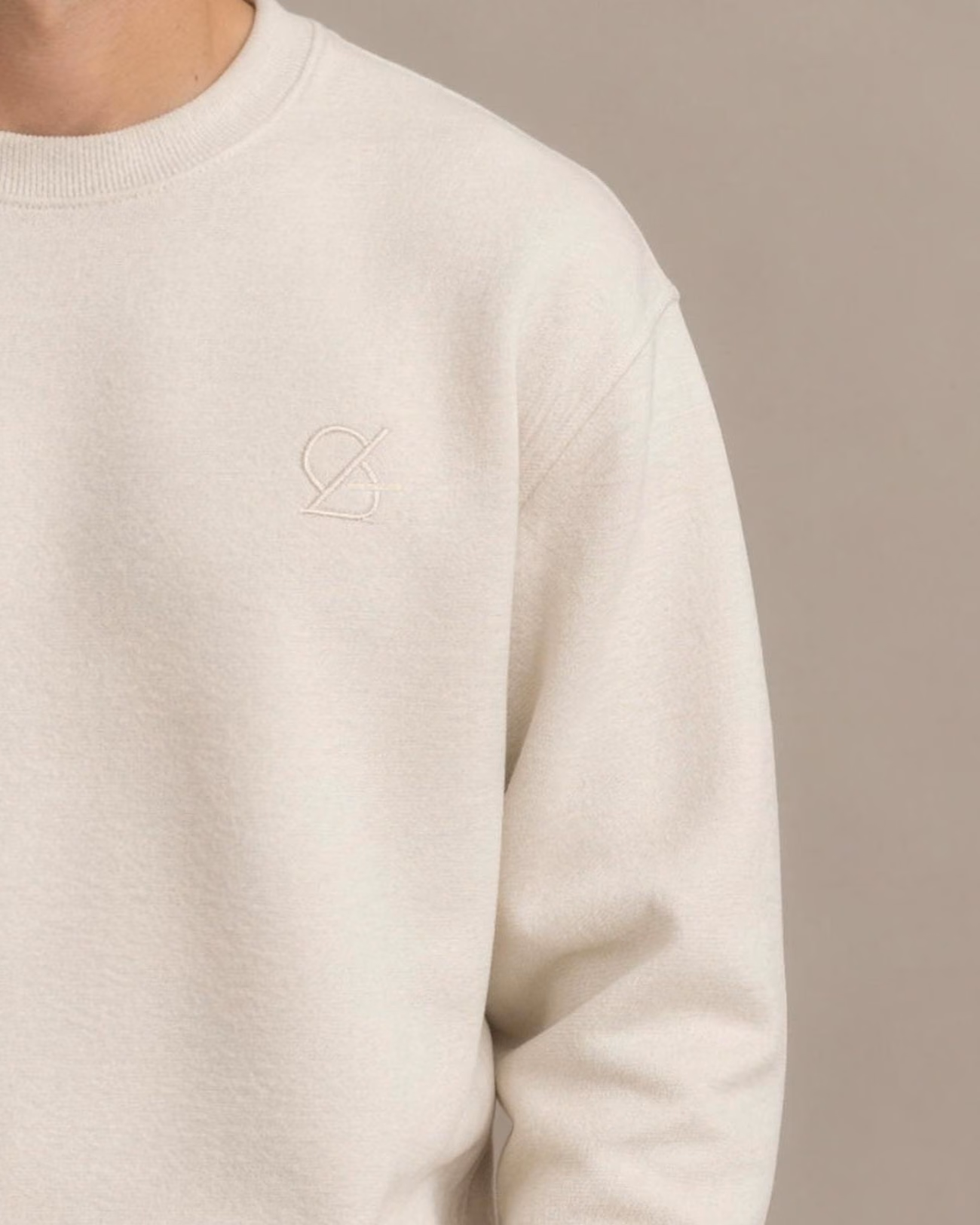 Close-up of the same image: Cream sweatshirt with a tone-on-tone embroidered emblem on the chest